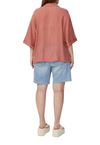Roestkleurig shirt met korte mouwen en een grove afwerking, gecombineerd met lichtblauwe denimshorts met een rafelige zoom. Neutrale platformsandalen.