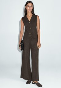 Gilet marron sur mesure avec des boutons dorés, associé à un pantalon à jambes larges. Tissu souple ; le mannequin tient un sac à main noir et porte des mocassins marron.