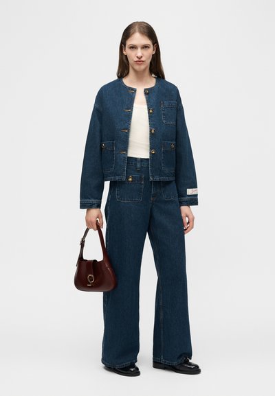 Γυναίκα που φοράει σκούρο μπλε τζιν μπουφάν και παντελόνι με φαρδιά πόδια, κρατώντας ένα καφέ δερμάτινο τσάντα στο αριστερό της χέρι, στέκεται μπροστά σε απλό φόντο.