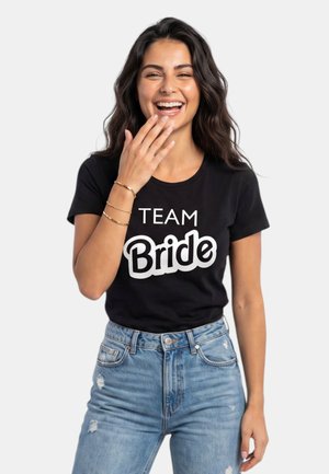 Mujer sonriendo con la mano cerca de la boca, lleva puesta una camiseta negra "Team Bride" y jeans azules rasgados sobre un fondo liso.