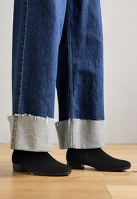 Schwarze Wildleder-Stiefeletten mit niedrigem Absatz, getragen zu weiten blauen Jeans aus Denim, die hellgraue Säume und ausgefranste Kanten aufweisen. Hintergrund aus Holzfußboden.