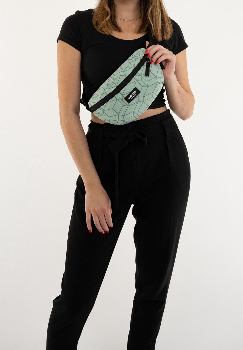 Femme portant un crop top noir et un pantalon, tenant un sac banane vert clair à motif géométrique croisé sur sa poitrine, avec des ongles rouges visibles.