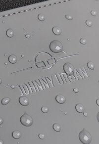 Szürke vízálló anyag, amelyen kiemelkedő betűk olvashatók: "JOHNNY URBAN", a felületen több vízcsepp található, hangsúlyozva a nedvességgel szembeni ellenállását.