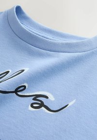 Tricou din bumbac de culoare bleu deschis, cu guler rotund, având un design cu text cursiv alb-negru pe față și o textură moale.