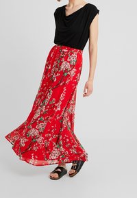 Jupe maxi florale rouge au design fluide, ornée de motifs de fleurs blanches et vertes. Associée à un haut drapé noir et des sandales noires.