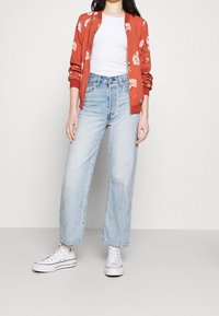 Veste bomber rouge à motif floral portée sur un T-shirt blanc, associée à un jean taille haute bleu clair et des baskets blanches.