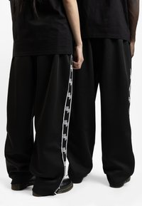 Pantalones negros de pierna ancha con franjas laterales blancas que presentan texto, tejido de textura fina y cremalleras cerca de los tobillos. Sin marcas visibles.