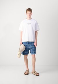 Camiseta blanca de algodón con el logo de Calvin Klein, pantalones cortos de cuadros azules, gorra beige y sandalias marrones con tiras ajustables.