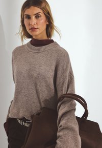 Heled pruun, ülelaia villasweater, millel on ribitud ümar kaelus. Kombineeritud tumeda pruuni nahkrepiga ja burgundi punase kõrge kaelusega pluusiga all.