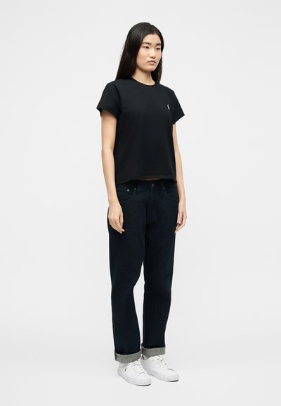 Νεαρή γυναίκα στέκεται, φορώντας μαύρο μπλουζάκι, σκούρο τζιν με γυρισμένα μπατζάκια και λευκά αθλητικά παπούτσια, μπροστά σε απλό ανοιχτό φόντο.