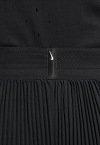 Musta urheilumekko, jossa on teksturoitu yläosa, verkkovyötärö ja laskostettu hame. Varustettu pienillä ilmanvaihtoaukoilla ja Nike-logolla varustetulla tunnisteella.