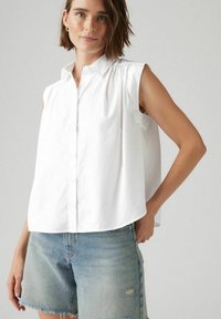 Vit ärmlös skjorta med knappar och strukturerad krage, mjukt tyg och avslappnad passform, kombinerad med slitna denimshorts.