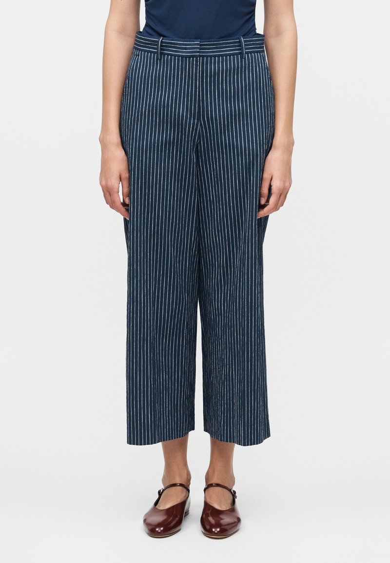 Femme portant un pantalon large court bleu marine à rayures verticales blanches et des chaussures Mary Jane marron foncé.