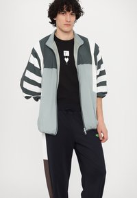 Let og zip-up jakke i blød grå med hvide og mørke grå striber. Bæres over en sort skjorte med et lille grafik. Sorte bukser med detaljer.