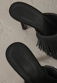 Mules à talons hauts en daim noir avec des accents de franges. La semelle intérieure présente des détails de marque en relief. Le talon robuste offre un support supplémentaire.
