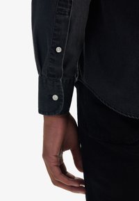Musta denimin paita napillisella hihalla, jossa on valkoinen nappi. Kangas näyttää teksturoidulta, siinä on pehmeää, tasalaatuista ommelta ja se on muotoiltu istuvaksi.