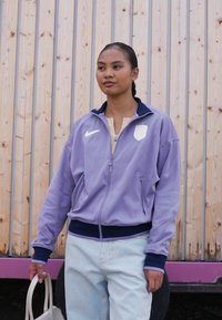 Lilla zip-up jakke med høy krage og marineblå detaljer, med Nike-logo og sidelommer, brukt over en lys topp og jeans.