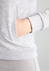 Hand som vilar i den främre fickan på en ljusgrå långärmad sweatshirt med ribbade ärmar och nederkant.