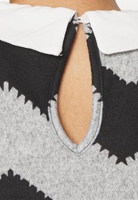 Gros plan sur un vêtement en maille gris et noir à motif zigzag avec un col blanc fermé par une boucle à bouton à l'arrière de l'encolure.