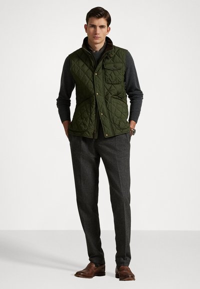 Zaļa siltināta veste ar brūniem akcentiem, augstu apkakli un kniedēm, saskaņota ar pelēkiem biksēm un brūnām apaviem. Gluda tekstūra un stilīgs dizains.