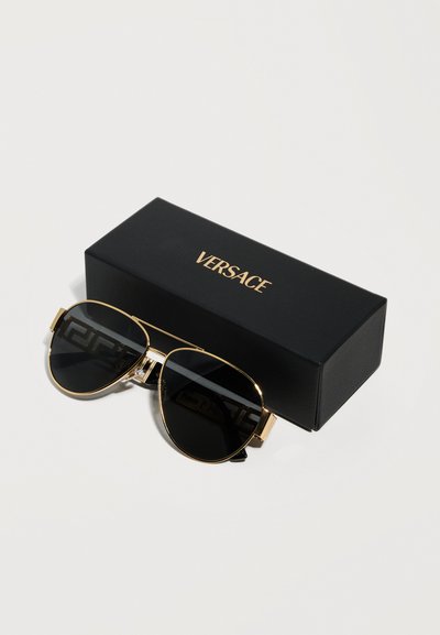 Γυαλιά ηλίου αεροπόρου με χρυσή κορνίζα και σκουρόχρωμους φακούς που ακουμπούν δίπλα σε μια μαύρη θήκη με το λογότυπο της Versace πάνω σε λευκή επιφάνεια.