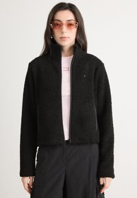 Sort fleecejakke med høy krage og glidelås. Lange ermer, myk tekstur, kombinert med en lyserosa skjorte under.