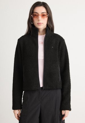 Black fleece jacket with a high collar and zip closure. Long sleeves, soft texture, paired with a light pink shirt underneath.