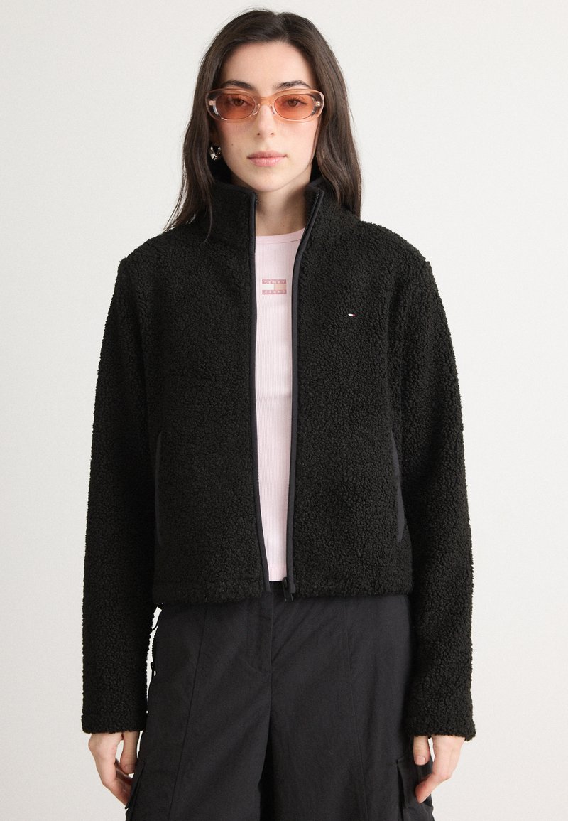 Sort fleecejakke med høy krage og glidelås. Lange ermer, myk tekstur, kombinert med en lyserosa skjorte under.