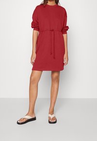 Robe rouge confortable en tissu doux, dotée d'une coupe décontractée, d'une taille à cordon et de manches trois-quarts, associée à des tongs simples.