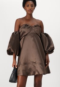 Barna selyem off-shoulder ruha puff ujjaival, sima textúrával és laza fazonnal. Egy kisméretű fekete táska van a kezében.