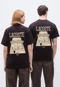 Τα μαύρα βαμβακερά T-shirts διαθέτουν ένα μεγάλο μπεζ γραφικό στην πλάτη με κείμενο και λογότυπα με θέμα το τένις, αναδεικνύοντας εμφανώς τη μάρκα Lacoste.