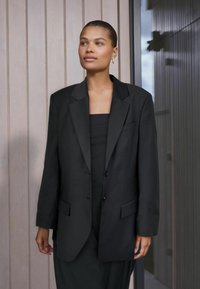 Černý propracovaný blejzr s jednou řadou knoflíků, klopovými reverzy, dvěma předními kapsami a hladkou, jemnou texturou. Styling přes černé tričko.