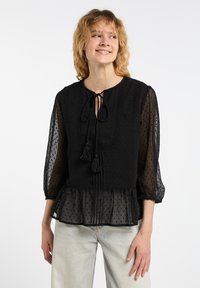 Sort bluse med gennemsigtige ærmer, prikket tekstur, blondekant og snøring med frynser ved halsudskæringen. Peplum-forneden tilføjer volumen.