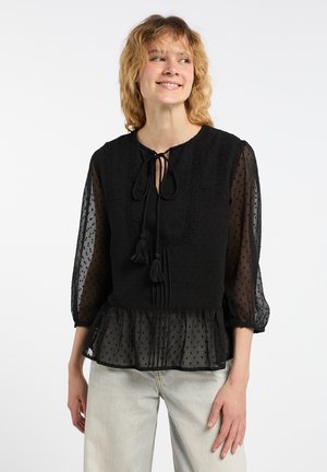 Blusa nera con maniche trasparenti, texture a pois, bordo in pizzo e una coulisse con nappine al collo. L'orlo a peplo aggiunge volume.