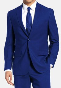 Veste de costume bleue avec revers en pointe, deux poches avant et fermeture à un seul bouton. Associée à une chemise blanche et une cravate bleue à pois.