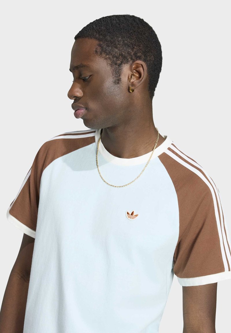 Junger Mann, der nach links schaut, trägt ein weiß-braunes Adidas T-Shirt mit Logo, eine Goldkette und einen goldenen Creolenohrring vor hellem Hintergrund.