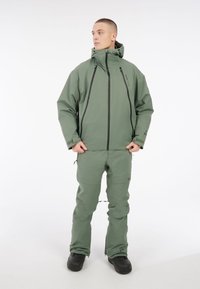 Muška dvodijelna odjeća za vanjske aktivnosti u maslinasto zelenoj boji, koja se sastoji od jakne s kapuljačom i odgovarajućih hlača s crnim patentnim zatvaračima i izdržljivom tkaninom.