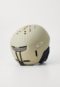 Casco de esquí beige con una superficie texturizada y orificios de ventilación. Presenta detalles en negro, correa ajustable y la marca "OAKLEY MOD3".