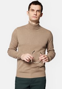 Beżowy sweter z golfem wykonany z gładkiego dzianinowego materiału, z prążkowanymi mankietami i dołem, noszony z ciemnozielonymi spodniami, trzymany w ręce w okularach.