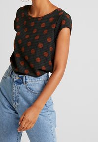 Haut noir avec des pois marron et un motif de grille texturé. Manches courtes et encolure ronde. Assorti avec un jean taille haute bleu clair.