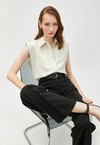 Weiße ärmellose Bluse mit Kragen, kombiniert mit schwarzen weit geschnittenen Hosen und einem strukturierten schwarzen Gürtel. Modell sitzt auf einem transparenten Stuhl.
