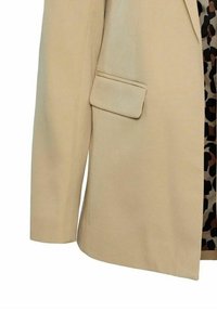 Blazer beige con una texture liscia, tasca frontale singola e spalle strutturate; tessuto con motivo visibile sotto in colori scuri.