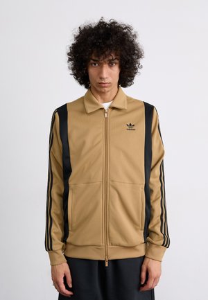 Kollane tõmbluku jaotusega jakk, mis on valmistatud tekstuursetest kangast, mustade külje triipudega, kolme musta triibuga varrukatel ja adidas logo rinnal.