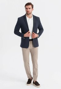 Man justerar en marinblå kavaj över en vit polotröja, bär beige byxor, svart bälte, klocka och mörkbruna mockaskor med tofsar.