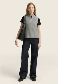 Szary, bezrękawnik z dzianiny z suwakiem, pod spodem czarny t-shirt, w połączeniu z szerokimi ciemnymi dżinsami i dużą czarną torbą typu tote.