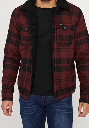 Mand iført en rød og sort ternet jakke med sort fleeceforing over en sort skjorte og blå jeans, stående med afslappede arme.