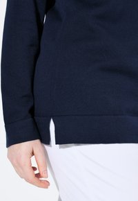 Tmavě modrý pletený svetr s dlouhými rukávy, s bočními rozparky na spodním lemu a texturovanou tkaninou. Detail rukávu a bočního prvku.