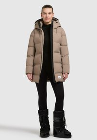 Beige, puffy jacket with a hood, featuring a black zipper and trim. Worn over black leggings and black snow boots. Simple design.