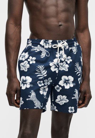 Mees torso, kes kannab tumelillaid ujumispükse valgete lille- ja leopardimustritega, millel on kummipael ja valge pael.