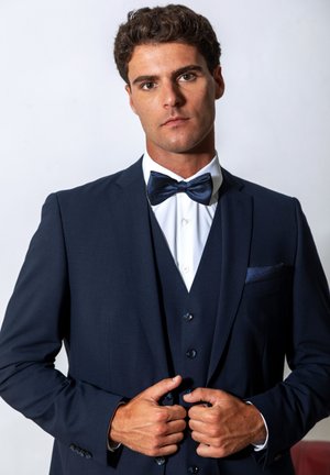Man adjusting navy blue suit jacket and vest, wearing white shirt and patterned blue bow tie against plain background.